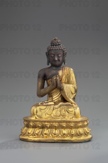 Shakyamuni Buddha, unknown sculptor, Himalayan, Sino-Tibetan style, Tibetan Buddhist art; Buddha ca. 560–480 BCE, ushnisha, lotus throne, mudras; 18th century, Tibet; sculpture, figurine; height 11 cm, width 7.7 cm., A seated statuette of a robed figure with hands held together in front, rendered in a combination of dark brown and gold tones; the figure has a textured hair bun and elongated earlobes, a serene facial expression, and detailed drapery on the robe; the base is ornate with carved petal-like patterns and is predominantly gold with some worn darker spots; the background is a smooth gray.