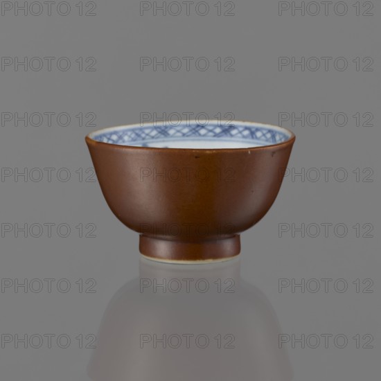 Alcohol bowl, Chinese art, Chinese culture, Qing 1644–1911, ceramics, blue-and-white porcelain, landscape motifs, 19th century, China, vessel, height 4.5 cm, diameter 7.8 cm, A small ceramic bowl with a smooth brown exterior and a glossy finish sits centered on a gray surface, showing a few subtle imperfections on its outer wall; the interior rim displays a decorative pattern in blue on a white background, and a narrow lighter band appears at the base where the bowl meets the surface, with a soft reflection of the bowl visible below.