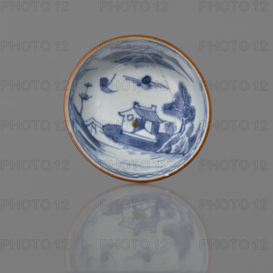 Alcohol bowl, Chinese, Qing dynasty 1644–1911, 19th century. Blue-and-white porcelain vessel with landscape motifs; height 4.5 cm, diameter 7.8 cm., A shallow circular ceramic dish with a brown rim sits against a gray background, showing a blue painted scene inside that includes a small building with a pitched roof, surrounding trees, a few clouds, and stylized landscape brushwork; the dish casts a soft, muted reflection below it that mirrors the blue-and-white interior design and brown edge.