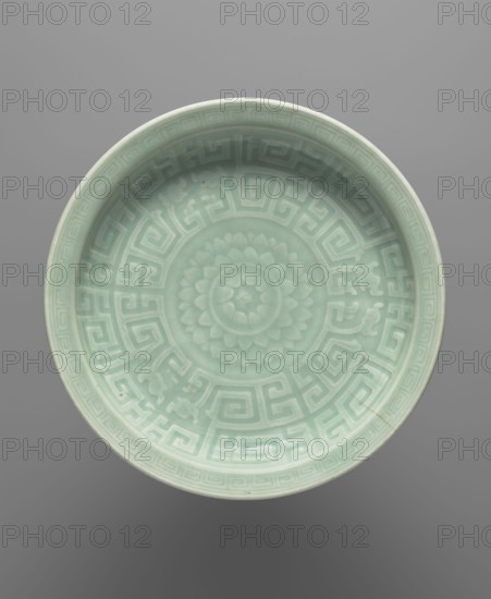 Plate - celadon, Chinese, Chinese art, lotuses, dragons iconography, ceramics, 18th century, China, vessel, height 5.4 cm, diameter 28.5 cm, A shallow round ceramic dish shown from above with a pale celadon green glaze featuring a raised central floral rosette surrounded by a geometric maze-like band and decorative corner motifs, with the rim and interior displaying subtle variations of light green and mint tones against a neutral gray background.