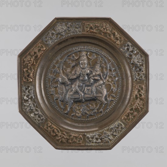 Plate, gift; Indian art; Indian culture; Hinduism, Shaivism; Shiva; Nandini; Parvati mythology; animals — antelopes, bulls; plants; ornaments; garlands; dedications; marriages; processions; shells; mounts; tiaras; crowns; family. On the rim an ornament with a Kirtymathy motif. 19th century, Southern India; vessel; height 0.9 cm; diameter 16.5 cm., An octagonal metal plate with a raised central circular medallion depicting a carved scene of human figures seated on an animal-like creature, surrounded by ornate foliage and animal motifs; the plate shows a mix of brown, dark bronze, aged copper, and hints of greenish patina across its surface, with darker recessed areas accentuating the carved details and lighter, warmer metallic highlights on the raised decorative borders.