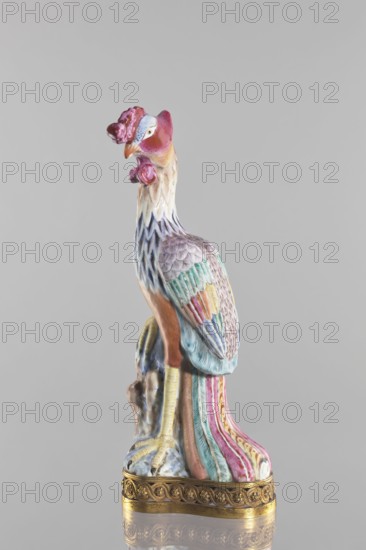 Bird, unknown creator; Chinese, Qing 1644–1911; porcelain figure of a bird pheasant/phoenix/fenghuang on a brown gilded openwork base with braided ornament and rosettes; colored porcelain; sculpture; 18th–19th century; height 40.4 cm, width 10.3 cm., A ceramic bird figurine in a standing pose on a decorative base, showing colors including pink, purple, blue, turquoise, yellow, orange, white, and gold; the bird has a textured feather surface with patterned wings and a long, multicolored tail, a curved beak, a crest and wattles on its head, and clawed feet positioned on a rock-like support, all set against a plain light gray background.