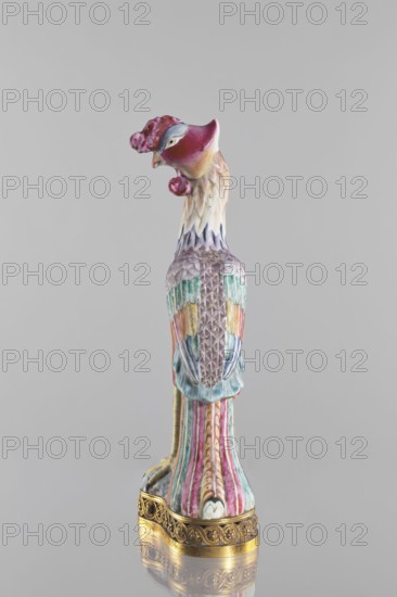 Bird, Chinese, Qing 1644–1911, 18th/19th century. Porcelain figure of a bird pheasant/phoenix, fenghuang iconography in colored porcelain on a brown gilded openwork base with braided ornament and rosettes. Sculpture; height 40.4 cm, width 10.3 cm., A ceramic bird sculpture standing upright on an ornate golden base, with a head turned to the side and a curved beak; the head and crest show shades of deep pink, magenta, and small areas of pale yellow, while the neck has cream and light beige tones blending into soft lavender. The body features a textured pattern of feathers in muted purple, teal, and turquoise with accents of pale yellow and coral, and the wings exhibit a mix of blue and green hues. The tail and lower body display vertical fluted stripes in pink, teal, and cream with subtle gold detailing, and the gilded base has intricate carved patterns in a warm metallic gold. The background is a uniform light gray.