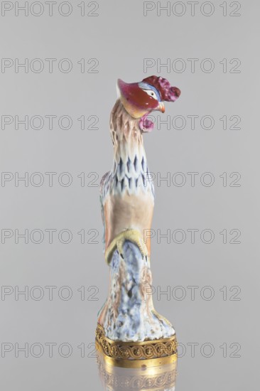 Bird, Chinese porcelain figure on a brown gilded openwork base with braided ornament and rosettes; colored porcelain depicting a bird pheasant/phoenix — fenghuang; Qing dynasty 1644–1911, 18th–19th century, China; sculpture; height 40.4 cm, width 10.3 cm., The image shows a tall ceramic bird figurine standing on an ornate gold-colored base; the bird has a predominantly cream and pale beige body with accents of white and blue on its neck feathers, a reddish-purple head with touches of pink and a small orange beak, and a pale yellow curved detail near its lower body; the lower portion of the figurine has mottled blue and white glaze with hints of tan and green, and the gold-colored base features decorative raised patterns.
