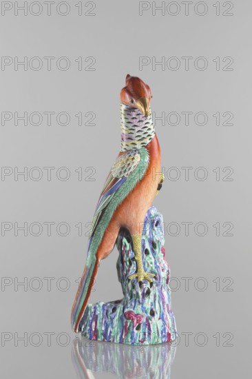 Pheasant figurine, Chinese, Qing dynasty 1644–1911, 19th century. Colored porcelain sculpture of a pheasant on rocks. Materials: ceramics, colored porcelain. Dimensions: height 49.5 cm, width 19.8 cm, depth 17.0 cm; weight 3.36 kg. Cultural attribution: Chinese; subject: birds pheasants., A colorful ceramic bird figurine perched on a textured base is shown against a plain light gray background, the bird displaying shades of orange and red on its head and body, a pale multicolored neck with small dark markings, and green and teal feathers along its wings accented with blue and purple tones; its beak and feet are golden yellow, and the base beneath it features a marbled mix of aqua, teal, purple, and pink with glossy highlights and small perforations, all reflected faintly on the smooth surface below.
