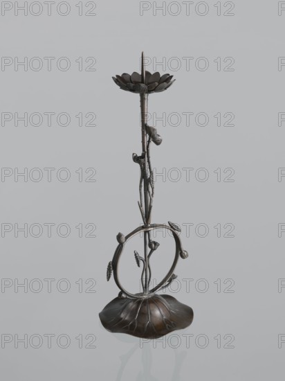 Candlestick shokudai, maker Takimoto, Iwami; Japanese; motifs: lotuses and leaves; Meiji period 1868–1912, 2nd half of the 19th century; Japan; candlestick; height 46.0 cm; provenance unknown., A dark metallic sculptural object resembling a tall stylized plant stands against a pale gray background; the piece has a petal-like base in dark brown with subtle lighter highlights, a slender stem rising from the center with textured nodes and small curled elements, a circular ring mid-height adorned with small bud-like protrusions and leaf forms, and a top element shaped like a shallow flower with rounded petals and a central pointed spike, all showing tones of dark bronze, brown, and black with metallic sheen.