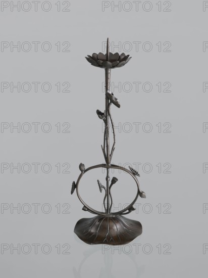 Candlestick shokudai, maker: Takimoto Iwami, Japanese; motifs: lotuses and leaves; Meiji period 1868–1912, 2nd half of the 19th century; Japan; medium: candlestick; height: 46.0 cm; provenance: unknown., A slender metallic sculpture with a dark brown and bronze finish stands against a pale gray background; the piece features a petal-like dish at the top with a pointed element rising from its center, a thin textured stem with several small bud-like forms attached, a circular open ring midway adorned with leaf and bud shapes, and a broad, curved base shaped like a stylized leaf that reflects slightly on the surface beneath.