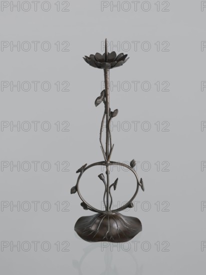 Candlestick shokudai, maker Takimoto, Iwami; Japanese; decorated with lotuses and leaves; style: Meiji period 1868–1912, 2nd half of the 19th century; material/technique: metal candlestick; provenance, height 46.0 cm, A dark bronze decorative stand shaped like a stylized flower with a long central stem rising from a broad, rounded base that resembles a leaf; a ring encircles the midsection with several small leaf and bud elements attached, and the top features a petal-like dish with a pointed spike; colors visible are dark bronze tones against a pale gray background.