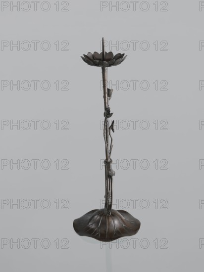 Candlestick shokudai, maker Takimoto, Iwami; Japanese; decorated with lotuses and leaves; Meiji period 1868–1912, 2nd half of the 19th century; Japan; candlestick; height 46.0 cm; provenance unknown., A slender metallic object with a dark bronze finish stands upright against a light gray background, featuring a rounded base shaped like a scalloped leaf with raised veins, a tall textured stem that resembles a plant stalk with a few small leaf and bud-like projections, and a shallow flower-shaped dish at the top with petal-like edges and a central pointed spike; visible colors include dark bronze, brownish tones, and the light gray of the background.