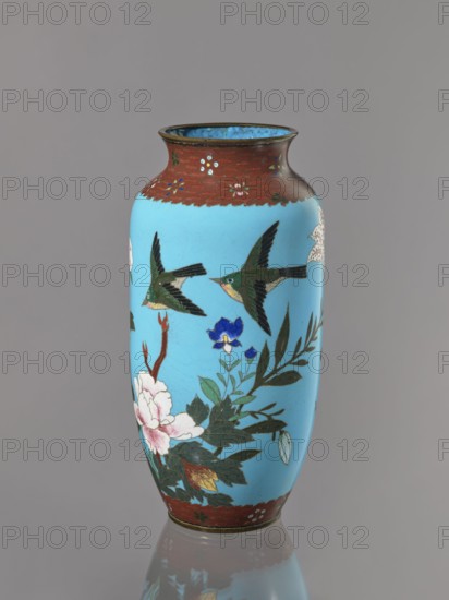 Vase, Cloisonné enamel on copper with brass rims at the spout and foot. Short, slightly flared neck. Decoration on a blue background: large motifs of rose bushes and lilies, small flowers, two butterflies and a bird kachôga. Japanese, Meiji period, ca. 1905–1908. Vessel; height 30.7 cm, diameter 10.6 cm., A tall cylindrical vase with a rounded shoulder and flared rim sits against a neutral gray background; the main body is a bright sky blue with painted motifs including a group of green and black birds in flight and floral arrangements featuring white and pink blossoms, a blue flower, and dark green leaves, with some reddish-brown branches. The neck and base areas have a band of mottled reddish-brown decorated with small floral accents, and the interior rim shows a darker blue. The vase casts a soft reflection on the surface below.
