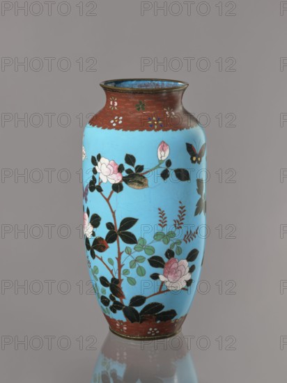 Vase. Cloisonné enamel on copper with brass rims at the spout and foot. Short, slightly flared neck. Decoration on a blue background: large motifs of rose bushes and lilies, small flowers, two butterflies and a bird kachôga. Japanese, Meiji period, ca. 1905–1908. Height 30.7 cm; diameter 10.6 cm., A tall ceramic vase with a glossy light blue body and a narrow neck finished in a reddish-brown band, decorated with painted floral branches, leaves, and blossoms in shades of pink, white, green, and dark green, with several black leaves and stems, a small painted butterfly with black and yellow wings, and additional small floral motifs on the reddish-brown rim and base; the vase casts a faint reflection on a smooth gray surface and is set against a plain gray background.