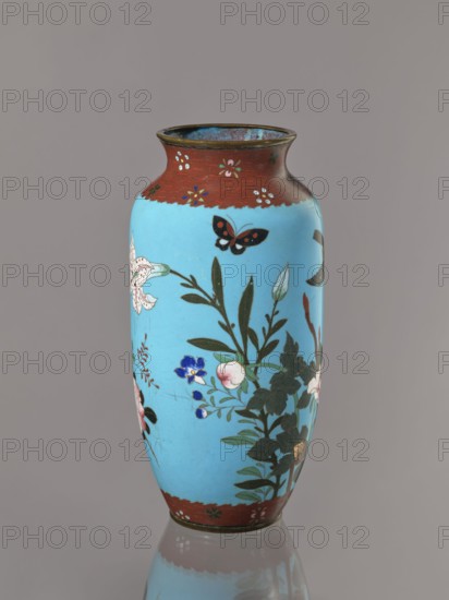 Vase, Cloisonné enamel on copper with brass rims at the spout and foot. Decoration: large roses and lilies, small flowers, two birds and a butterfly on a blue background kachôga. Short, slightly flared neck. Meiji period, ca. 1905–1908, Japan. Vessel; height 30.8 cm, diameter 10.6 cm., A tall vase with a glossy light blue body and a reddish-brown rim and base, decorated with painted floral branches, leaves, and blossoms in colors including white, pink, blue, green, and black, and showing a black and red butterfly near the upper portion; the vase sits against a neutral gray background and its lower portion is reflected on the surface beneath it.