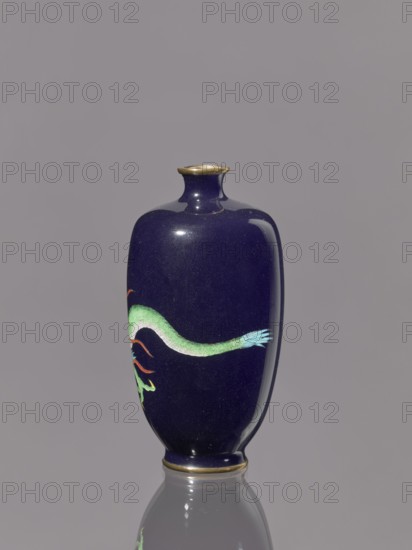 Vase, Meiji period 1868–1912, late 19th–early 20th century. Cloisonné enamel on silver. Vase on a low, ring-shaped foot with a short, thin neck slightly flared at the spout; decorated with cloisonné enamel featuring dragons Japanese iconography. Japan. Vessel. Height 11.8 cm; diameter 6.1 cm., A glossy, deep blue ceramic vase with a narrow neck and slightly flared rim, the rim and base edges accented in a metallic gold color, standing on a reflective surface that mirrors its lower portion; the vase features a painted sinuous form in shades of green with a pale green to turquoise tail and a few red-orange curling accents against a plain gray background.