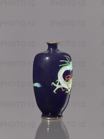 Vase, Meiji period 1868–1912, Japan. Cloisonné enamel on silver. Vase on a low, ring-shaped foot with a short, thin neck slightly flared at the spout. Decoration includes dragons iconography. Vessel; height 11.8 cm, diameter 6.1 cm., A glossy vase with a deep navy surface stands against a plain gray background, featuring a painted dragon motif in shades of green, pale green, turquoise, and touches of red and orange, with a small bright green and turquoise accent on the opposite side; the vase has a narrow neck and a slightly flared rim with a thin metallic gold band at the top and bottom, and its surface reflects light, showing highlights and a faint reflection on the gray surface beneath.