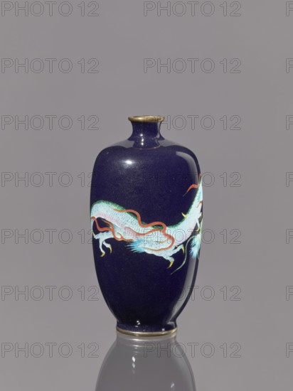 Vase, repossession; gift; dragons iconography; Japanese art; Japanese dragon motif; Meiji 1868–1912; cloisonné enamel. Vase on a low, ring-shaped foot with a short, thin neck slightly flared at the spout. Decoration with cloisonné enamel on silver. 19th/20th century Meiji period, Japan. Vessel. Height 11.8 cm, diameter 6.1 cm., A glossy, deep blue ceramic vase with a narrow neck and rounded body sits upright against a plain gray background, featuring a decorative stylized dragon-like motif in pale aqua, white, orange, green, yellow, and light blue that wraps around its midsection; the vase has a thin metallic rim at the mouth and base and casts a faint reflection on the surface beneath it.
