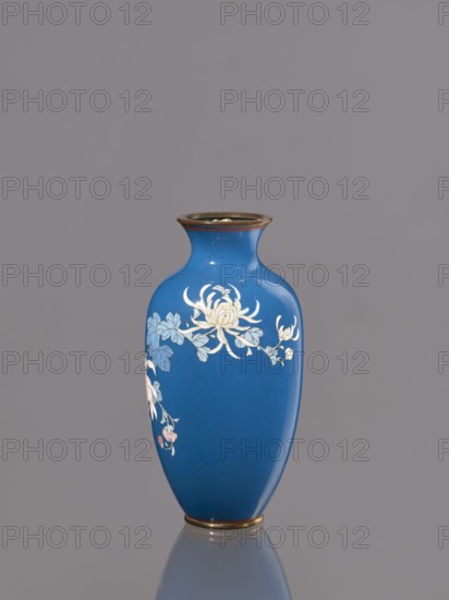 Vase with chrysanthemums, ca. 1900 Meiji 23, Japan, Nagoya; cloisonné enamel vessel; height 18.0 cm, diameter 9.0 cm., A blue vase with a narrow neck and flared rim sits against a gray background, showing a glossy surface and a reflective base. The vase features a floral design with pale yellow and white blossoms and blue-green leaves, and small touches of pink on some buds. The rim and base have a metallic-looking band in a warm brownish-gold tone.
