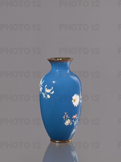 Vase with chrysanthemums, ca. 1900 Meiji 23, Japan, Nagoya; cloisonné enamel vessel, height 18.0 cm, diameter 9.0 cm; subject: chrysanthemums/flowers., A glossy blue vase with a narrow neck and flared rim sits centered against a gray background, showing decorative floral motifs in white, pink, light blue, and pale yellow on its curved body, with a thin metallic band of bronze or gold around the rim and base and a subtle reflection of the vase visible on the surface below.