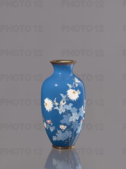 Vase with chrysanthemums, ca. 1900 Meiji 23, Japan, Nagoya; cloisonné enamel vessel, height 18.0 cm, diameter 9.0 cm; Japanese Meiji period; chrysanthemums and flowers iconography., A blue vase with a narrow neck and flared rim stands against a plain gray background, decorated with white and pale yellow flowers and blue leaves, with touches of pink on some smaller blossoms and a thin metallic band at the top and base, its glossy surface producing subtle highlights and a faint reflection on the surface beneath.
