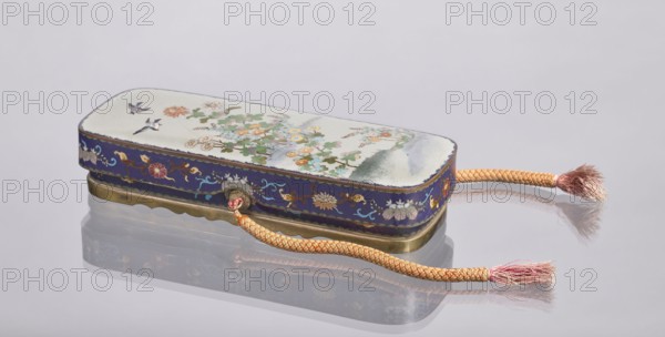 Letter box fubako with lid, Japanese art, Japanese culture, Meiji 1868–1912, cloisonné enamel box with birds and chrysanthemums, late 19th century Meiji period, Japan; container; height 4.6 cm, width 23.0 cm, depth 8.8 cm., A rectangular decorative box with rounded corners rests on a reflective surface, showing a mirrored reflection beneath; the box has a pale top surface painted with floral motifs and a few small birds, featuring colors such as white, green, orange, pink, yellow, and blue, and a blue band around the sides decorated with floral and scroll designs in gold, pink, white, and turquoise; the base has a metallic brass-colored trim. A braided cord in shades of peach, orange, and pink is attached to the box and extends outward, ending in frayed tassels in pink and beige. The background is a smooth, light gray color.