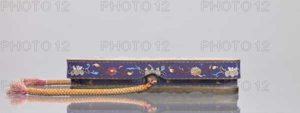 Letter box fubako with lid, Japanese, Meiji 1868–1912, cloisonné enamel, decorated with birds and chrysanthemums, late 19th century, Japan; container; height 4.6 cm, width 23.0 cm, depth 8.8 cm., A low, rectangular decorative box with a scalloped brass base and a deep blue exterior decorated with floral motifs in colors including white, pink, yellow, green, red, and light blue sits on a reflective surface, accompanied by a braided rope handle in shades of yellow and orange ending in a frayed tassel showing pink and beige tones; the background is a pale gray.