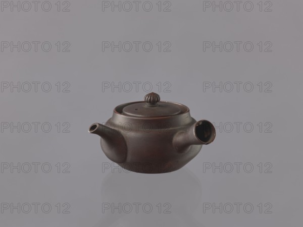 Teapot kyûsu, ceramics, Japanese art, teapots, Japanese culture, openwork, floral ornaments. Bulky teapot with a funnel-shaped handle decorated with an openwork floral rosette. Unglazed stoneware, thin shell. 19th century, Japan, vessel; height 6 cm, diameter 8.7 cm., A small round teapot with a low profile and a flared rim sits centered against a plain background; the teapot is a deep brown with subtle reddish undertones and a slightly glossy finish, featuring a short curved spout on one side and a tubular handle-like spout on the opposite side, a flat lid topped with a ridged knob, and soft highlights and shadows that reveal its smooth, rounded form, with its reflection faintly visible beneath it.