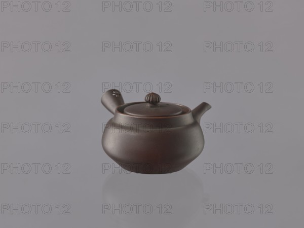 Teapot kyûsu, ceramics, Japanese art, teapots, Japanese culture, openwork, floral ornaments. Bulky teapot with a funnel-shaped handle decorated with an openwork floral rosette. Unglazed stoneware, thin shell. 19th century, Japan. Vessel; height 6 cm, diameter 8.7 cm., A small round teapot with a lid and a side handle sits against a plain background; the teapot is dark brown with subtle reddish-brown highlights, the background is gray, and there is a faint reflection of the teapot below it.