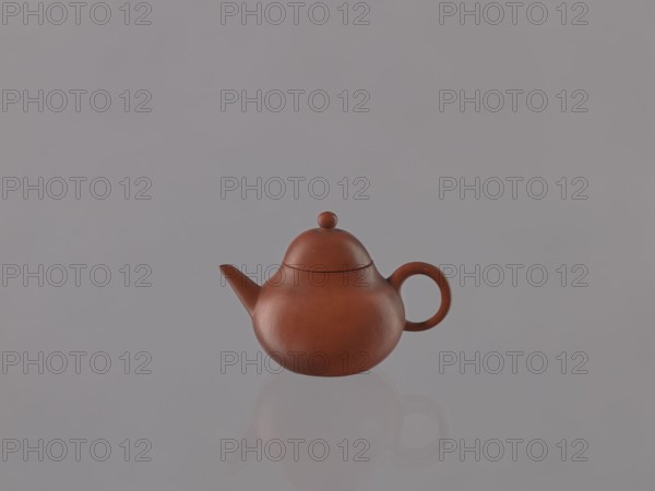 Teapot with lid, Chinese art, Chinese culture, Qing 1644–1911, ceramics, teapots, Yixing style, tea accessories, red stoneware, 19th century, China, Yixing, Jiangsu, vessel, height 6.1 cm, diameter 9 cm, A small round teapot in a reddish-brown clay color sits centered against a smooth light gray background, with a rounded lid topped by a small knob and a curved handle on one side and a short spout on the other, and a soft reflection of the teapot visible below it.