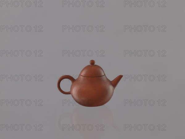 Teapot with lid; maker, ; Chinese art; Chinese culture; Qing dynasty 1644–1911; red stoneware Yixing teapot; 19th century, China; Yixing, Jiangsu; tea vessel; height 6.1 cm, diameter 9 cm., A brown rounded teapot with a circular handle and a short angled spout sits centered against a smooth pale gray background, its rounded lid topped with a small knob and a soft reflection visible below; colors present include brown and pale gray.