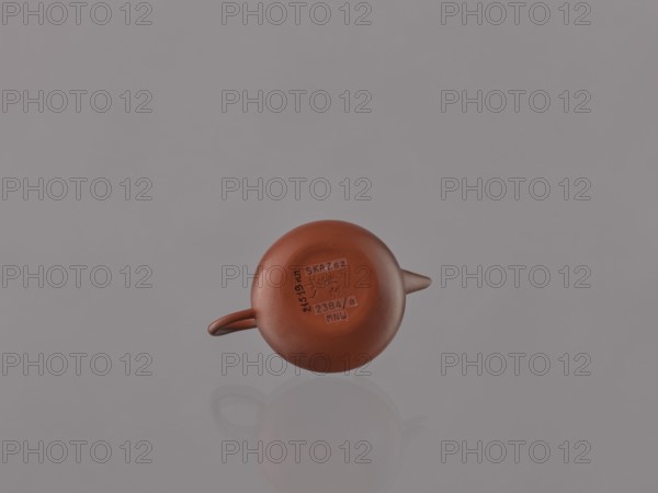 Teapot with lid;, ; Chinese art; Chinese culture; Qing dynasty 1644–1911; Yixing-style red stoneware teapot; 19th century; China, Yixing, Jiangsu; tea accessory vessel; height 6.1 cm, diameter 9 cm., A small round reddish-brown teapot with a short spout and a small handle sits centered against a smooth pale gray background, showing a subtle reflection beneath it; the surface of the teapot is matte and warm in tone, and the image contains shades of reddish-brown, pale gray, and a hint of darker brown.