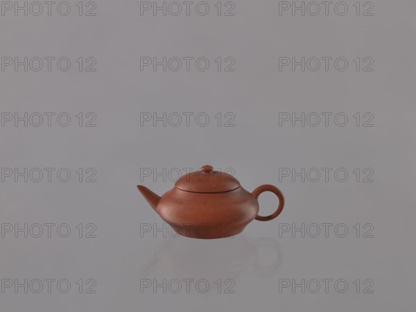 Teapot with lid, Chinese, Qing dynasty 1644–1911, 19th century. Yixing red stoneware teapot, Yixing style, Jiangsu province, China. Dimensions: height 4.0 cm, width 9.1 cm. Subjects/functions: teapot, tea accessory., A small reddish-brown teapot with a rounded body, short spout, and circular handle sits centered on a smooth reflective surface, casting a faint reflection; the background and surface are muted gray, with the teapot showing subtle highlights and darker reddish-brown shading.