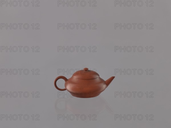 Teapot with lid; maker,  noted; Chinese, Qing dynasty 1644–1911; Yixing red stoneware teapot; 19th century; Yixing, Jiangsu, China; tea accessory vessel; height 4.0 cm, width 9.1 cm., A small reddish-brown teapot with a rounded body, a short curved spout, a circular handle, and a domed lid sits centered on a smooth reflective surface, casting a soft reflection beneath it against a muted gray background with subtle variations in tone.