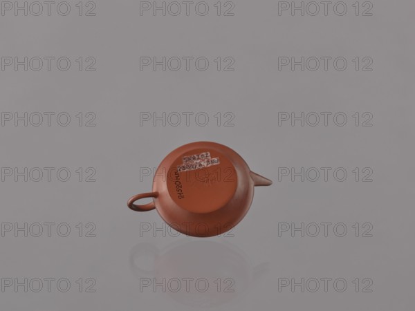 Teapot with lid; maker, Glezmer, Stanislaw 1853–1916 associated; Chinese; Qing dynasty 1644–1911; 19th century; Yixing-style red stoneware teapot; Yixing, Jiangsu, China; tea accessory vessel; height 4.0 cm, width 9.1 cm, A small reddish-brown rounded teapot viewed from above, showing its handle on one side and a short spout on the opposite side, resting on a smooth reflective gray surface that creates a faint reflection; the background is a uniform light gray.