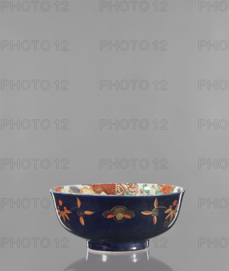 Bowl, Japanese, 2nd half of the 19th century, Arita. Colored porcelain vessel decorated with lotuses, fantasy animals, butterflies, flowers, and peonies; fo iconography. Height 10.8 cm, diameter 24.5 cm., A shallow bowl sits on a reflective surface against a plain gray background, the bowl exterior a deep navy blue with decorative floral motifs in orange, gold, and red, and the interior rim showing multicolored patterns including red, green, and blue.