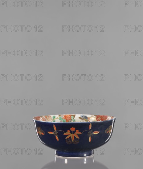 Bowl, Japanese, Arita, 2nd half of the 19th century. Colored porcelain vessel decorated with lotuses, fantasy animals, butterflies, flowers, and peonies; fo iconography. Height 10.8 cm, diameter 24.5 cm., A dark blue ceramic bowl with a glossy finish sits on a reflective surface. The bowl has an outward-flaring rim and a short foot. The exterior shows a pattern of stylized floral motifs in orange and gold with thin gold outlines and spiral accents. The interior rim reveals a multicolored decorative band with visible greens, reds, blues, and gold against a light background. The overall visible colors include dark blue, orange, gold, green, red, blue, and a light cream.