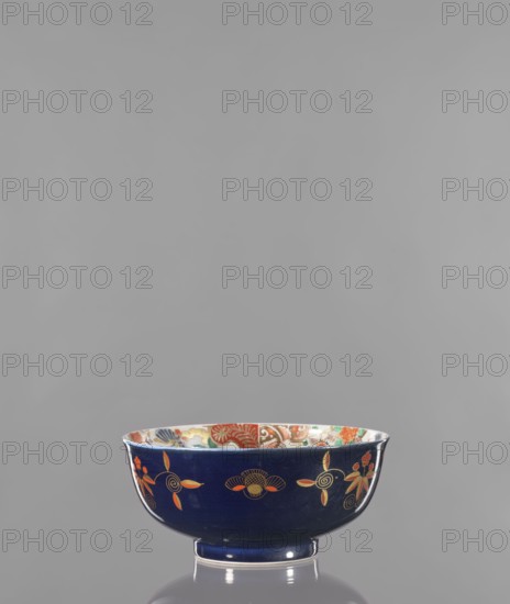Bowl, ; colored porcelain; decorated with flowers, lotuses, and fantastic animals fo; Japanese art, Arita, Japan; 2nd half of the 19th century; vessel; height 9.8 cm, diameter 21.9 cm., A ceramic bowl sits on a reflective surface against a plain gray background, the bowl exterior a deep navy blue decorated with a pattern of floral and spiral motifs in orange, gold, and pale yellow, while the interior rim shows a colorful painted band with shades of red, green, orange, blue, and white; the bowl has a short foot in matching navy blue and a glossy finish.