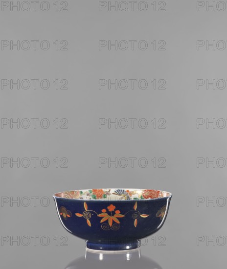 Bowl, colored porcelain, ceramics; decorated with flowers, lotuses, and fantastic animals fo iconography; Japanese art, Arita, Japan; 2nd half of the 19th century; vessel, height 9.8 cm, diameter 21.9 cm., The image shows a ceramic bowl with a deep blue exterior decorated with orange and gold floral motifs and spiral accents, and a richly patterned interior featuring colors including orange, green, and blue; the bowl sits on a small circular foot and rests on a reflective surface against a plain grey background.