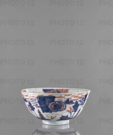 Bowl, Japanese, Edo 1603–1868, ca. 1680–1720. Colored porcelain vessel, height 12.4 cm, diameter 25.8 cm. Decoration: on the outside a stylized landscape with pagodas; on the inside large ornamental rosettes and flowered branches. Motifs include pagodas, plant ornaments, flowers, and a rosette., A porcelain bowl with a rounded shape and a short foot, decorated on its exterior with painted motifs in blue and orange-red on a white background, showing floral and abstract cloud-like patterns and a round medallion design, with several blue painted areas and orange-red detailing, sitting on a reflective surface against a neutral gray backdrop.