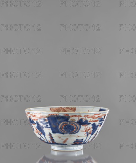 Bowl, Japanese, Edo 1603–1868, ceramics, colored porcelain; exterior decorated with a stylized landscape with pagodas; interior with large ornamental rosettes and flowered branches; ca. 1680–1720, Japan; vessel; height 12.4 cm, diameter 25.8 cm, A porcelain bowl with a white base is pictured against a plain gray background; the bowl features decorative painted motifs in deep blue and reddish-orange arranged around its exterior and rim, including rounded medallion-like shapes, cloud-like forms, stylized tree or plant shapes, and architectural or pagoda-like elements, with a glossy finish that reflects light and a short foot supporting the body.