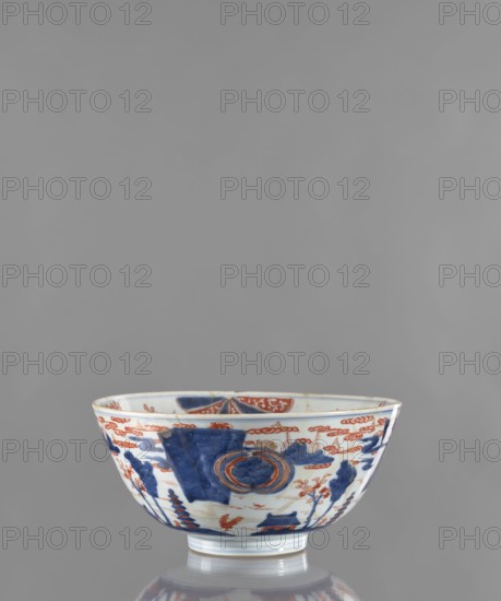 Bowl; maker,  noted. Japanese, Edo period ca. 1680–1720. Colored porcelain vessel, height 12.4 cm, diameter 25.8 cm. Decoration: outside shows a stylized landscape with pagodas; inside features large ornamental rosettes and flowered branches. Subjects: pagodas, plant ornaments, flowers, rosette., A shallow ceramic bowl with a rounded foot, shown against a plain gray background, decorated with a pattern in blue and reddish-orange on a white base; the exterior shows stylized landscape motifs including trees and architectural forms in blue, accented with reddish-orange cloud-like bands and floral elements, with a circular medallion motif in blue and reddish-orange on one side; the rim and interior edge have repeated decorative bands in the same colors, and the bowl sits on a reflective surface that shows a soft mirrored image.