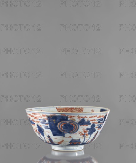 Bowl, Japanese, Edo 1603–1868, ceramics, colored porcelain. Decoration: on the outside a stylized landscape with pagodas; on the inside large ornamental rosettes and flowered branches. ca. 1680–1720, Japan. Vessel; height 12.4 cm, diameter 25.8 cm., A ceramic bowl with a rounded profile and a short foot sits against a gray background, decorated with hand-painted motifs in blue and orange-red; the exterior shows a large blue panel with circular blue-and-orange medallion designs, stylized blue foliage and cloud-like patterns, and scattered orange-red floral and decorative elements, while the rim and interior edge display additional orange-red accents; the bowl rests on a reflective surface that shows a faint reflection of its blue and white foot.