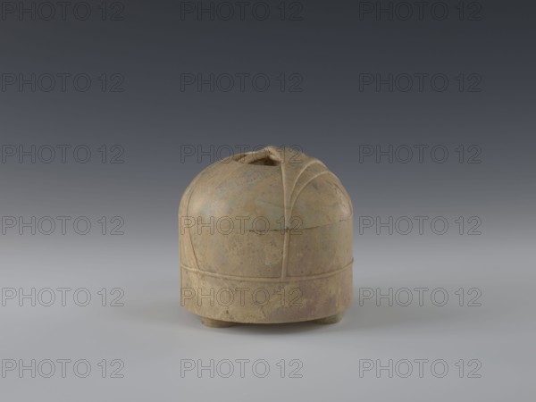 Tin with lid;, repossession Kraków removed. Chinese art; Chinese culture; Qing dynasty 1644–1911; 19th century; China place of origin?; yurts subject; container; height 12.8 cm; diameter 13.1 cm, A rounded, lidded ceramic vessel sits on a smooth surface, with a dome-shaped top and a cylindrical body raised on a few short feet; the lid has carved, petal-like ridges converging toward a small central opening. The surface shows a mottled, weathered texture with patches of tan, beige, muted brown, and pale green, and faint areas of darker brown and gray. The background transitions from a darker gray at the top to a lighter gray near the surface, and soft, diffused lighting casts subtle shadows beneath the object.