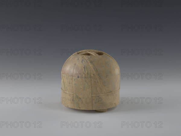 Tin with lid, gift, Chinese art, Chinese culture, Qing 1644–1911, yurts, 19th century, China, container; height 12.8 cm, diameter 13.1 cm, A rounded, dome-like object sits centered on a smooth surface against a gradient background that transitions from dark gray at the top to lighter gray near the bottom. The object is made of a mottled stone with a mix of tan, beige, brown, and hints of green-blue veining. Its surface shows carved lines and shallow grooves forming curved and vertical bands around the form, and there is a shallow recessed opening at the top. The base rests on a small circular foot, and the object casts a soft shadow on the pale gray surface.