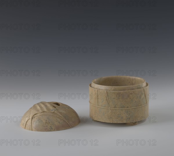 Tin with lid, Chinese, Qing 1644–1911, 19th century, possibly from China; container depicting yurts; height 12.8 cm, diameter 13.1 cm., A small round lidded container sits on a smooth surface with the lid placed beside it; both pieces appear made of a mottled stone showing shades of brown, tan, and muted green with subtle darker flecks, the container having a cylindrical form with carved horizontal and diagonal bands and a slightly recessed rim, and the lid having a domed shape with carved flowing ridges and a central oval opening, all set against a gradient background that transitions from dark gray at the top to lighter gray near the surface.