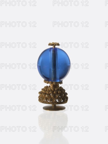 Button for a semi-official cap of a third-ranking official, blue glass ball mounted on a pin with a dome decorated with rosettes, 19th century Qing China, Chinese art; officials' emblem; jewelry/clothing accessory; height 6.6 cm, diameter 3.1 cm., A decorative object with a transparent blue spherical element mounted on an ornate metallic base is shown against a pale background; the blue sphere is glossy and reflects light, with a visible central rod running through it, and the metallic base and small top finial are detailed with clustered bead-like embellishments and filigree work in a gold tone, casting a soft reflection on the surface below.