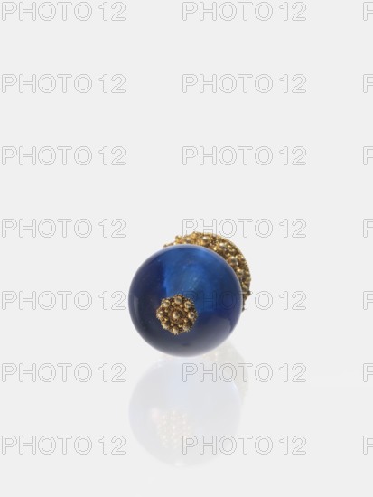 Button for a semi-official cap of a third-ranking official, Chinese art, Chinese culture, Qing 1644–1911, officials, emblems. Button composed of a blue glass ball mounted on a pin with a dome decorated with rosettes. 19th century, China. Jewelry, clothing; height 6.6 cm, diameter 3.1 cm., A single round blue glossy sphere with a small ornate gold floral cap attached to one side, resting on a reflective white surface that shows a faint mirror image; the visible colors are deep blue, gold, white, and subtle lighter blue highlights.