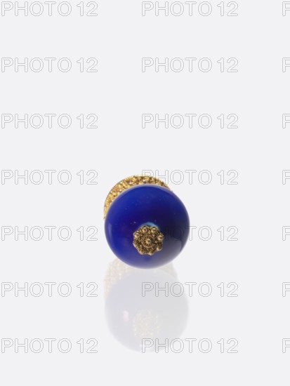 Button for a semi-official cap of a fourth-ranking official, Gift; 19th century, China, Qing 1644–1911. Chinese art; officials; emblems. Button composed of a blue-navy glass ball mounted on a pin with a dome decorated with rosettes. Jewelry/clothing. Height 6.6 cm; diameter 3.1 cm., A single round object with a glossy deep blue surface and a decorative metallic gold floral element at its center rests on a pale reflective surface, showing a soft reflection beneath; behind it a textured gold backing is partially visible. The visible colors are deep blue, gold, and pale gray-white.