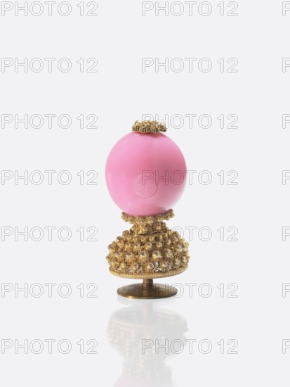 A button for a semi-official cap of an official between the seventh and ninth rank, Chinese art, Chinese culture, Qing 1644–1911, officials, emblems. A button composed of a glass opaque pink glass ball mounted on a pin with a dome decorated with rosettes; the ball is engraved with the character shou. 19th century, China; jewelry, clothing; height 6.0 cm, diameter 2.8 cm., A pink spherical object sits atop an ornate gold-colored stand; the sphere is smooth and glossy with a faint circular indentation visible, and the stand features intricate clustered floral-like gold detailing with a textured base and a small round gold pedestal, all reflected softly on the glossy white surface beneath. The visible colors are pink, gold, white, and a hint of brown on the pedestal.