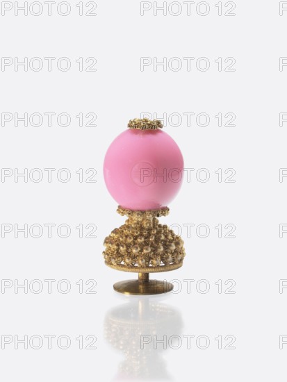 A button for a semi-official cap of an official between the seventh and ninth rank;, Qing dynasty 1644–1911, 19th century, China. A button composed of an opaque pink glass ball mounted on a pin with a dome decorated with rosettes; the ball is engraved with the character shou. Function: cap button for officials; materials: glass and metal. Height 6.0 cm; diameter 2.8 cm., A decorative object consisting of a smooth pink spherical element mounted on an ornate metallic base; the base is golden in color with a textured, beaded floral pattern and a round flat foot, and there is a small golden decorative collar at the top of the pink sphere; the scene has a pale gray background and a faint reflection of the object on the surface below.