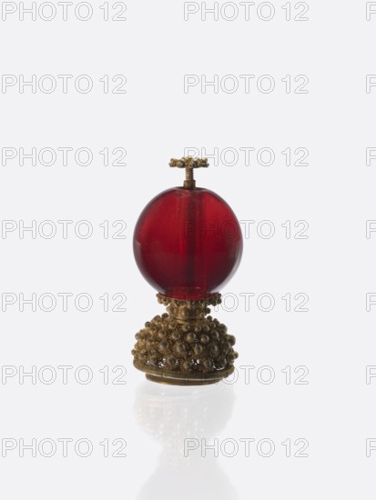 A button for a semi-official cap of a first-ranking official;, Chinese; Qing dynasty 1644–1911. Glass transparent ruby glass ball mounted on a pin with a dome decorated with rosettes. 19th century. Function: clothing ornament for officials; style: Chinese art. Height 6.6 cm; diameter 3.0 cm., A small decorative object sits centered against a pale background, consisting of a glossy deep red spherical element mounted on an ornate metallic base and topped with a small decorative metallic finial; the base and finial are a warm antique gold color with intricate beaded and floral-like textured detailing, while the red sphere is smooth and translucent with subtle highlights and internal variations in tone; the pale surface beneath the object produces a soft reflection showing muted gold and red hues.