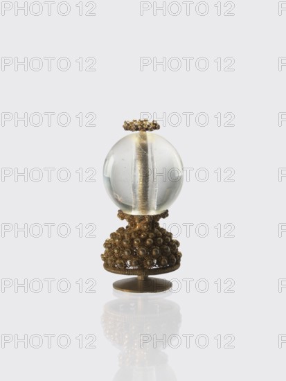 Button for a semi-official cap of a fifth-ranking official, Chinese, Qing dynasty 1644–1911. Button composed of a transparent glass ball mounted on a pin with a dome decorated with rosettes. 19th century. Materials: glass and metal. Function: cap button for an official. Dimensions: height 6.2 cm; diameter 2.9 cm., An ornate object sits against a pale gray background, consisting of a clear spherical glass orb perched on a decorative metallic base; the glass is transparent with subtle internal reflections and a central vertical element visible inside, and the metal base is a warm golden-bronze color with an intricate clustered beaded texture and floral-like motifs, topped by a small ring of matching beaded metal and standing on a round flat bronze pedestal that casts a soft mirrored reflection below.